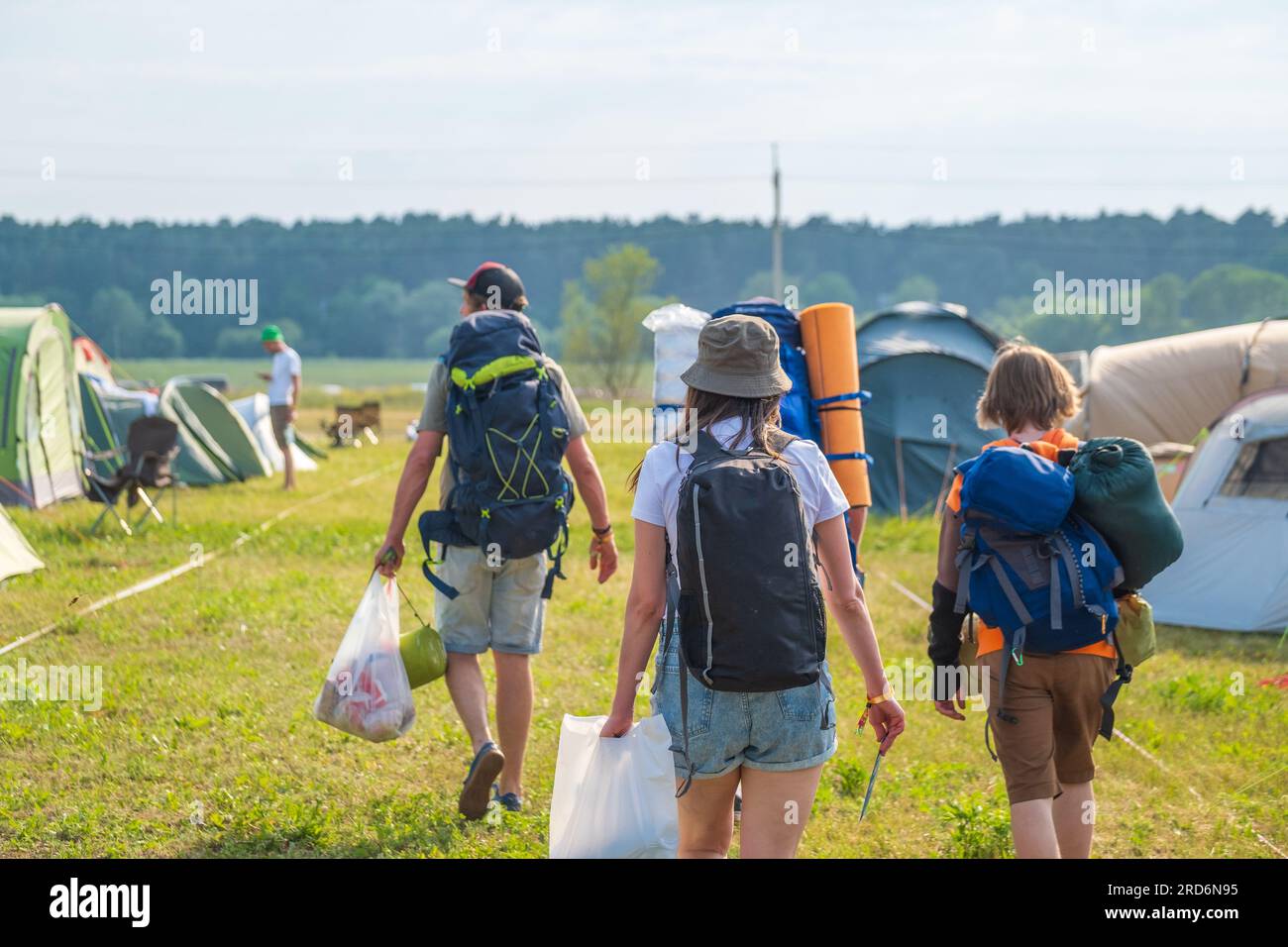 Vista posteriore di turisti irriconoscibili con zaini che trasportano borse mentre cammini lungo le tende da campeggio su prato erboso durante l'evento Foto Stock