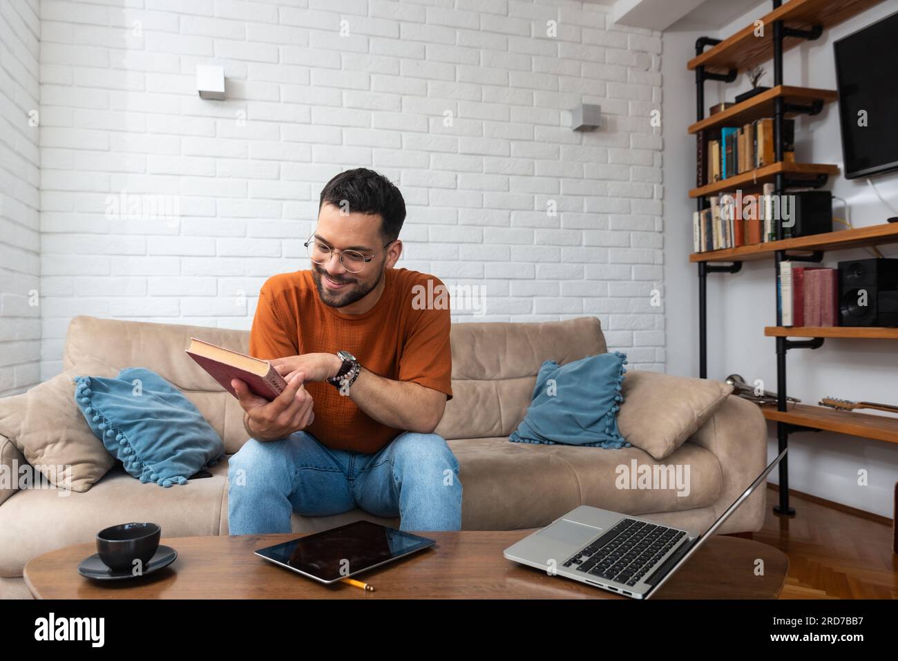Sorridente ragazzo millenario trascorri una serata intima in salotto seduto su un comodo divano, leggi il libro di carta preferito, bevi tè caldo. Il giovane lettore ha piacere enjo Foto Stock