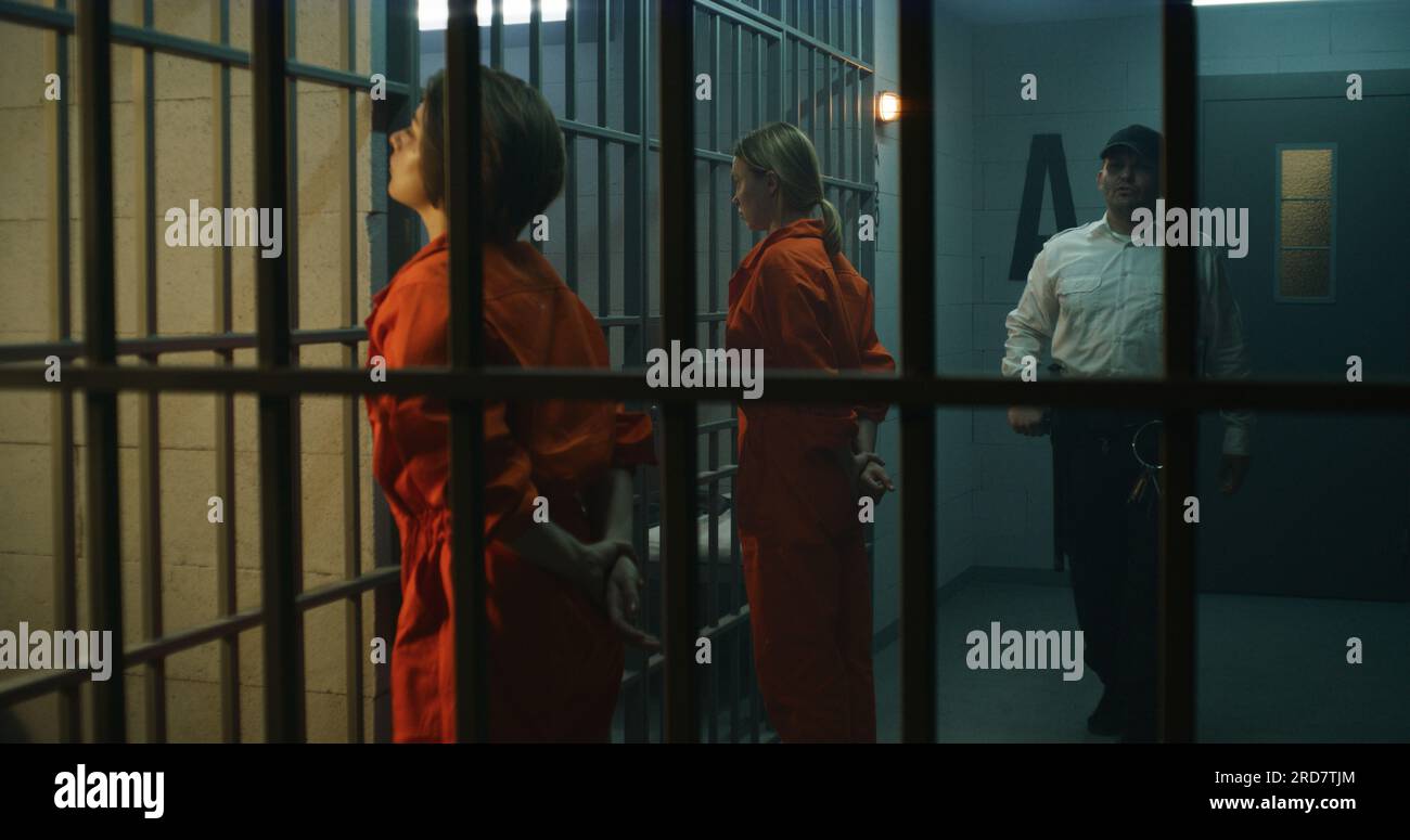 Due prigionieri femminili, detenuti in uniforme arancione, si trovano di fronte alle barre di metallo di fronte alle celle della prigione. L'agente di prigione cammina, guarda le donne criminali in prigione. Centro di detenzione o centro di correzione. Foto Stock