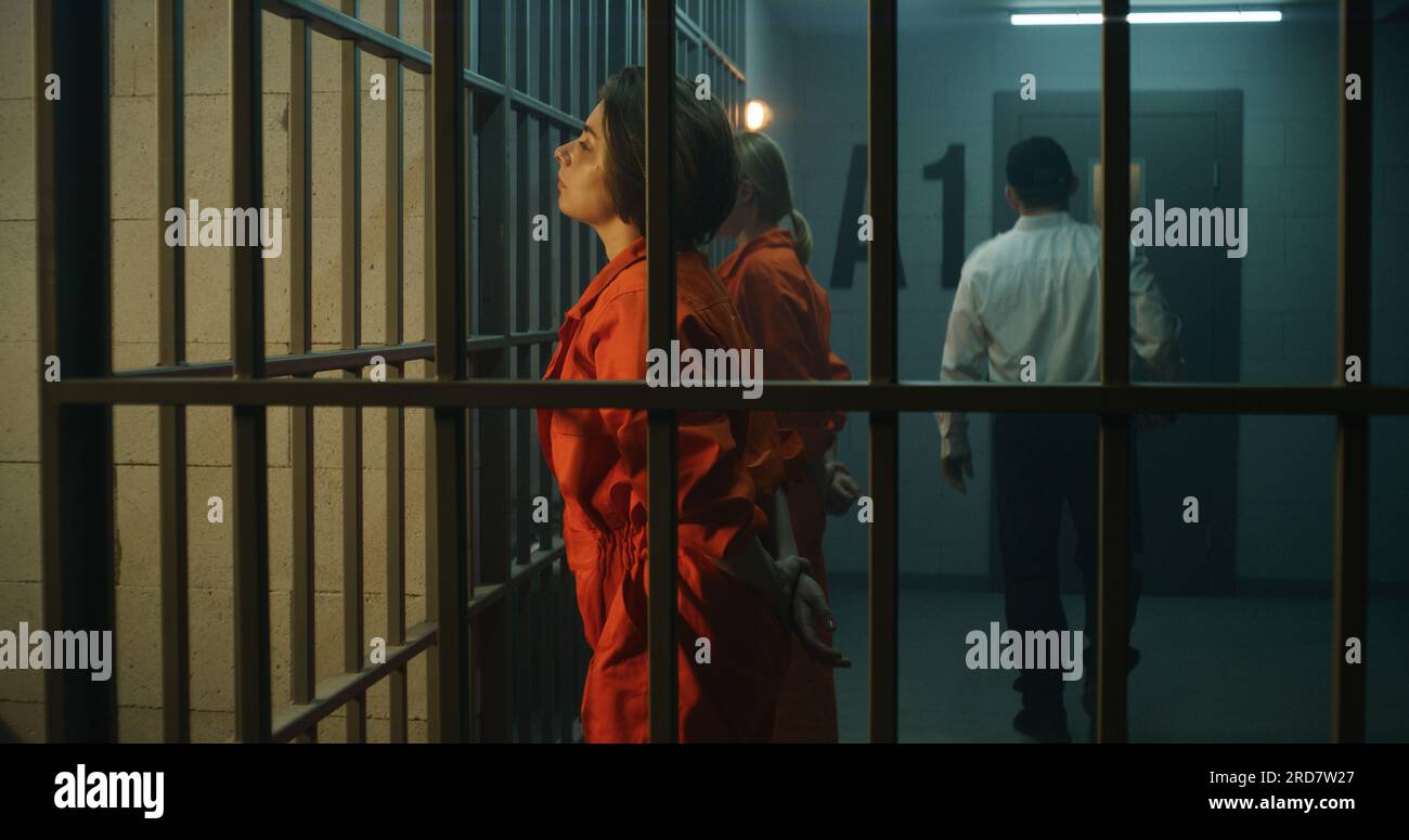 Due prigionieri femminili, detenuti in uniforme arancione, si trovano di fronte alle barre di metallo di fronte alle celle della prigione. L'agente di prigione cammina, guarda le donne criminali in prigione. Centro di detenzione o centro di correzione. Foto Stock