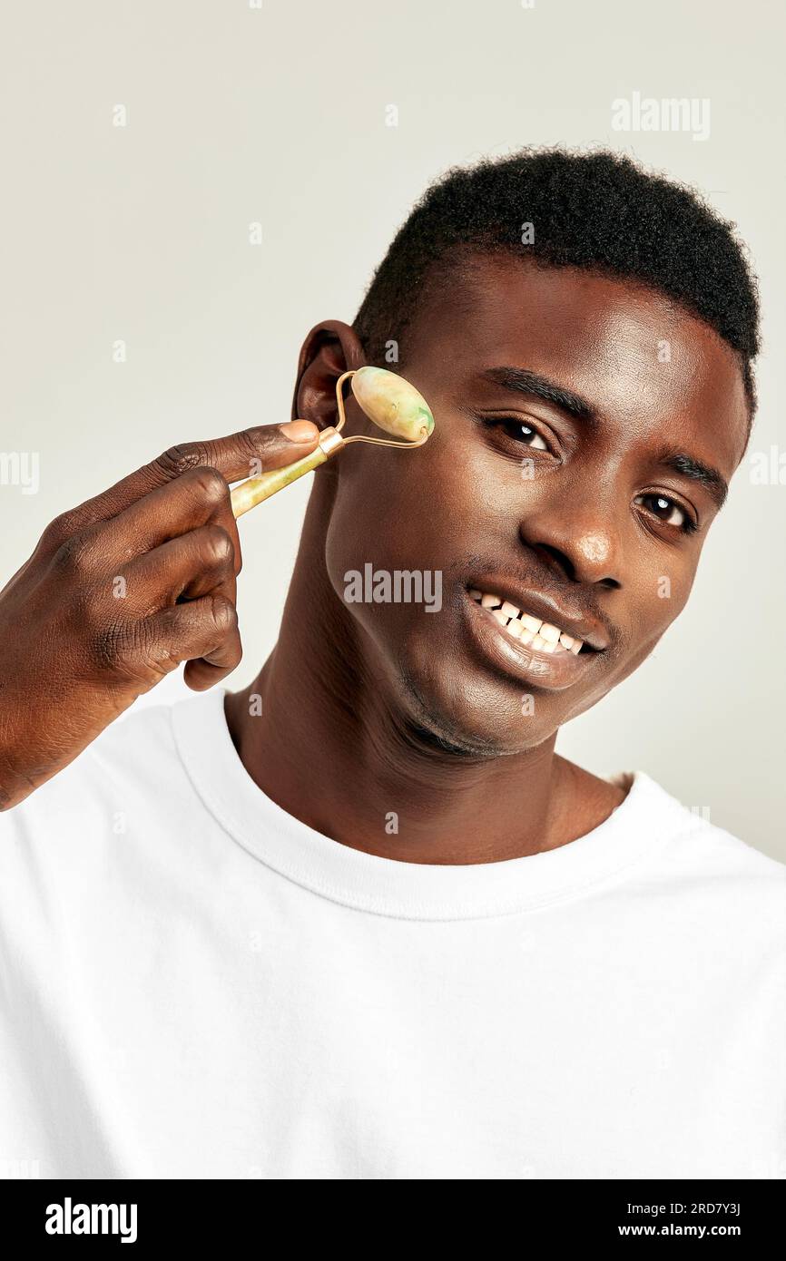 Un giovane ragazzo nero che usa il rullo facciale di degonfiore di giada per mantenere la sua pelle morbida e liscia. L'uomo millenario afroamericano mantiene la sua bea naturale Foto Stock