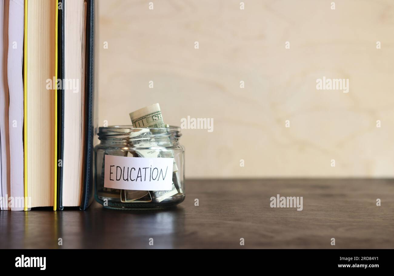 Vaso di vetro con denaro vicino a una pila di libri. Salvadanaio con dollari per l'istruzione su un tavolo di legno. Risparmia denaro per i futuri studi universitari. copia spac Foto Stock