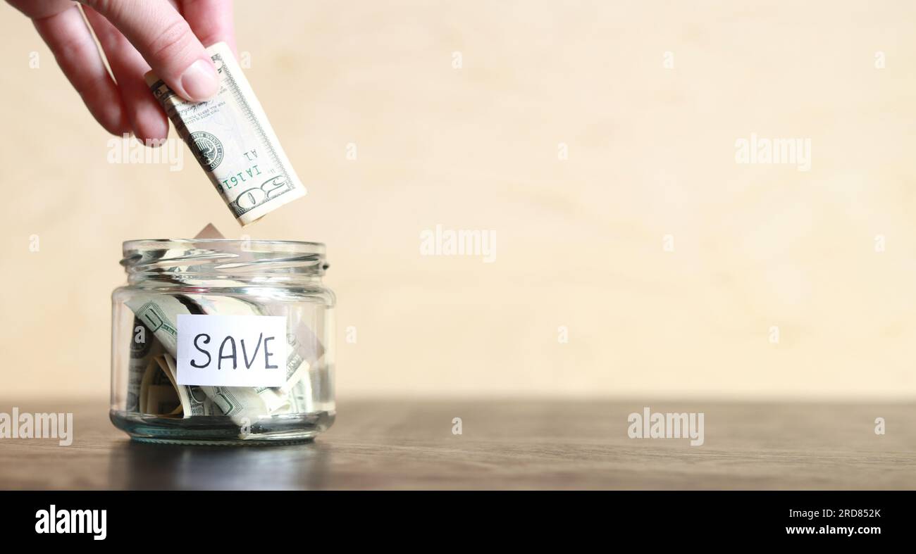 Risparmia denaro per il futuro. La mano di una donna mette una banconota da un dollaro in un barattolo di risparmi. Vaso di vetro con dollari su un tavolo di legno. Salvadanaio con banconote clo Foto Stock