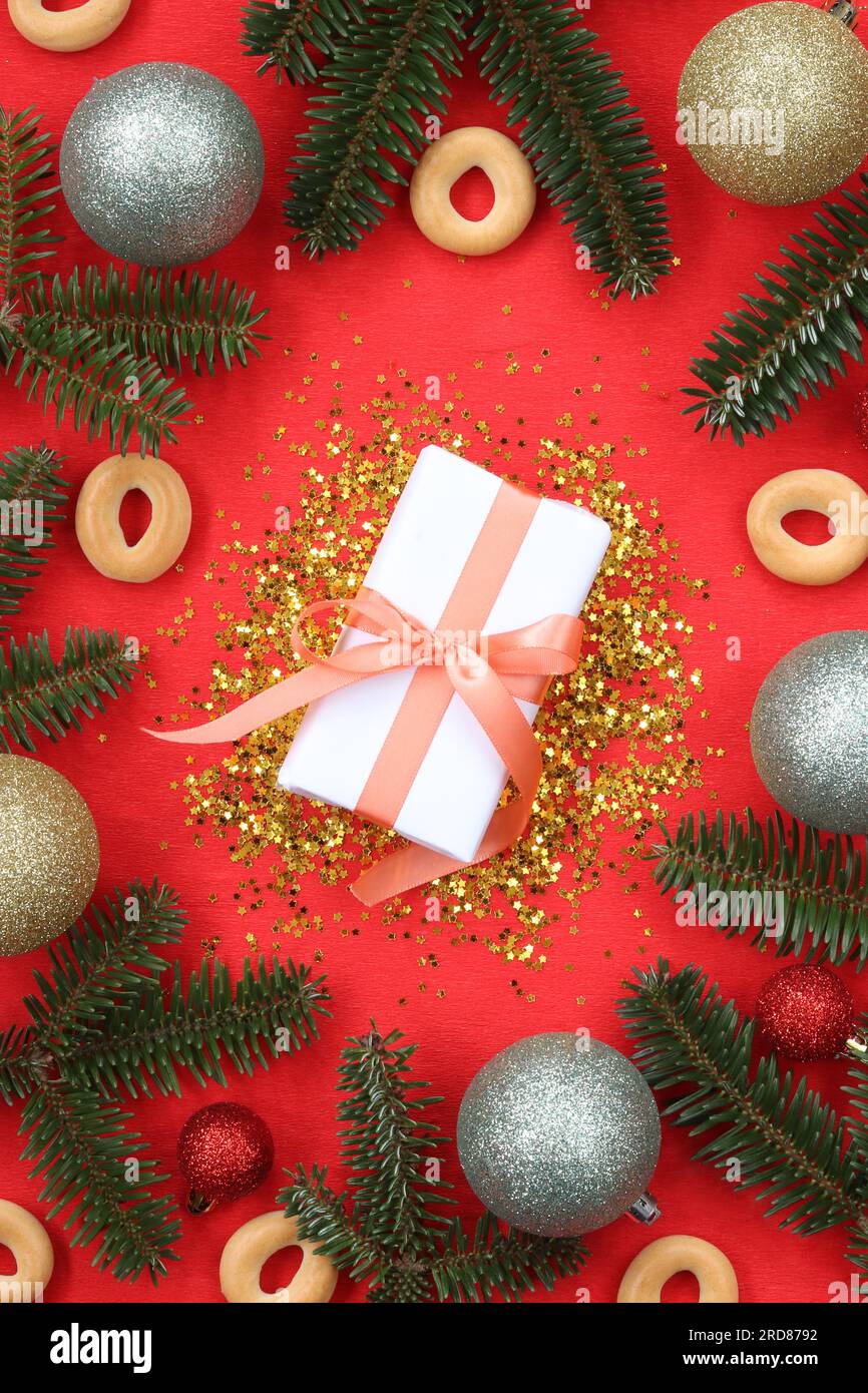 Regalo di Capodanno su sfondo rosso circondato da rami di abete. Composizione di paillettes dorate, decorazioni natalizie, rami e confezione regalo con fiocco Foto Stock