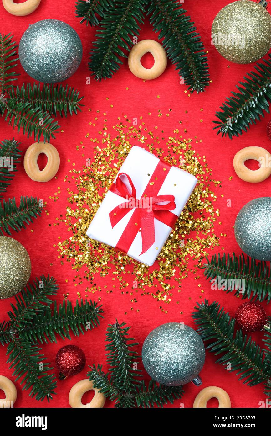 Regalo di Capodanno su sfondo rosso circondato da rami di abete. Composizione di paillettes dorate, decorazioni natalizie, rami e confezione regalo con fiocco Foto Stock