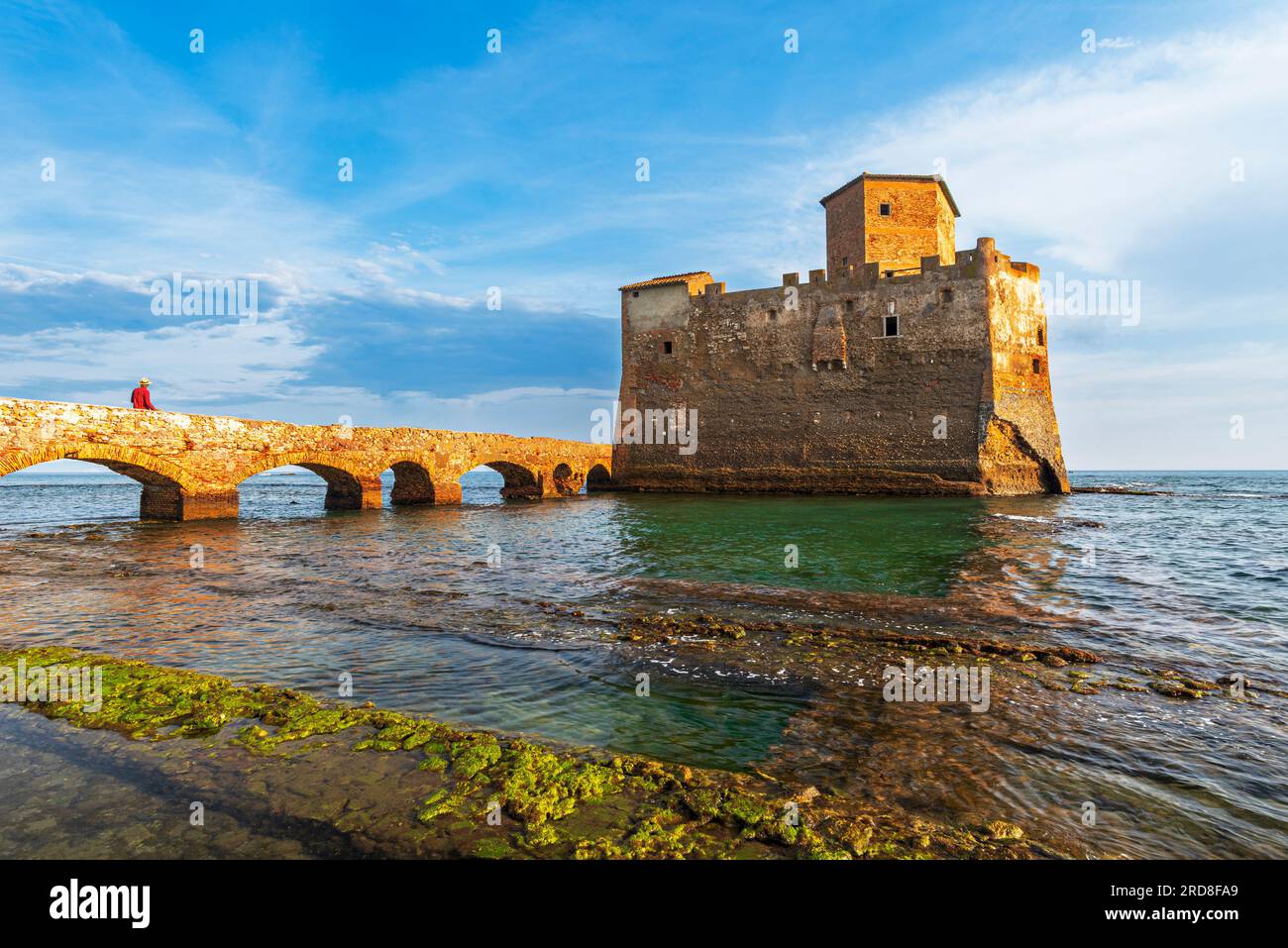 L'uomo sul ponte cammina fino al castello fortificato di Torre Astura, sulle acque del Mar Tirreno, costruito sulle rovine di una villa romana, ora del tramonto Foto Stock