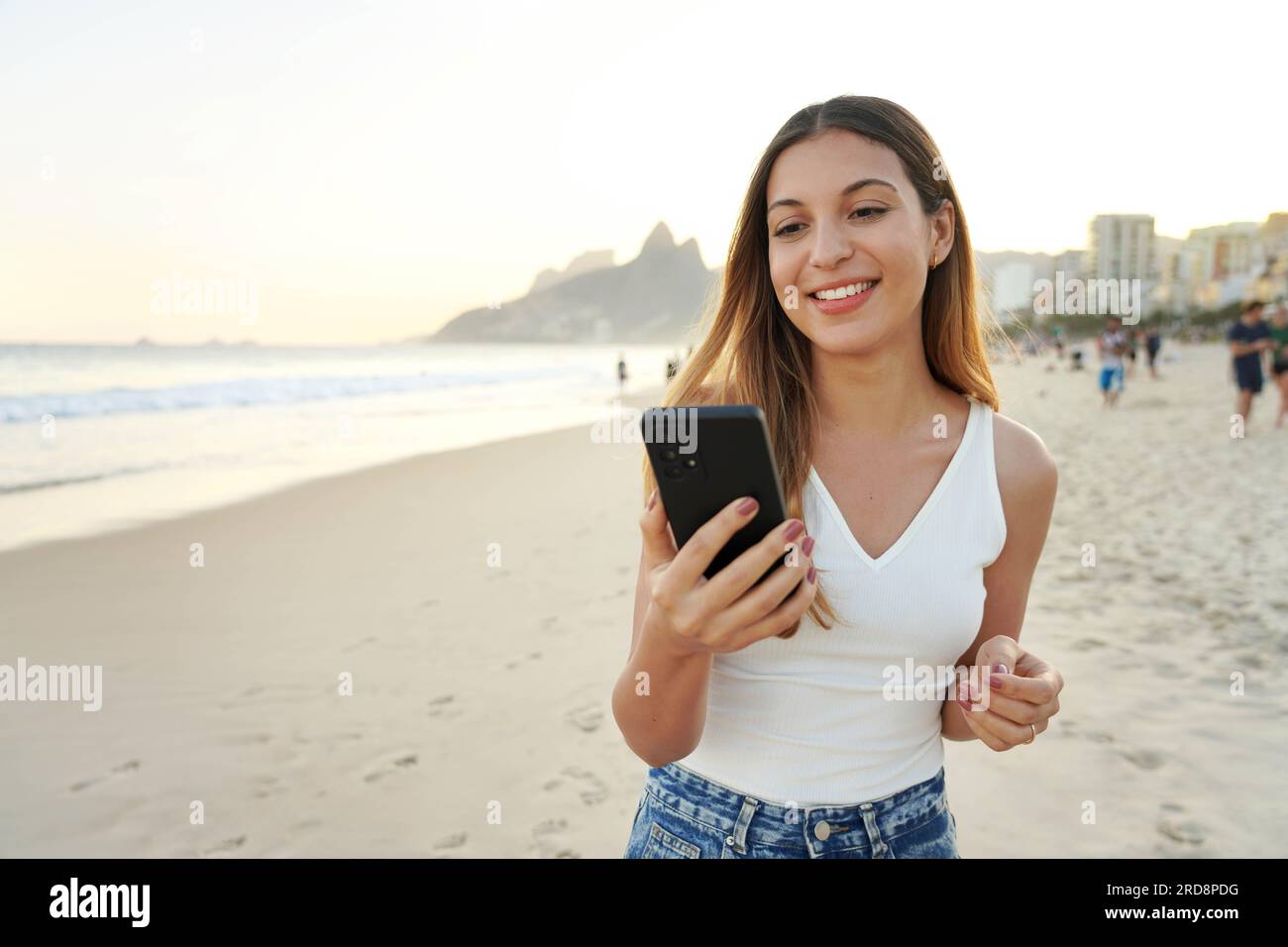 Ritratto di una giovane donna brasiliana sorridente sulla spiaggia di Ipanema che tiene in mano e guarda il suo smartphone, Rio de Janeiro, Brasile Foto Stock