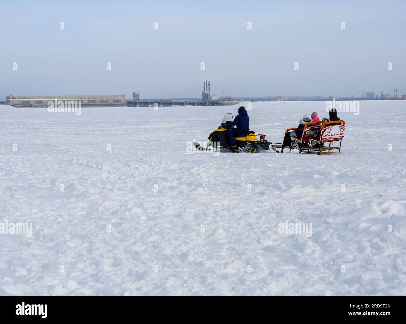 Una motoslitta trasporta una slitta con persone su un lago ghiacciato coperto di neve, con un'area industriale della città all'orizzonte Foto Stock