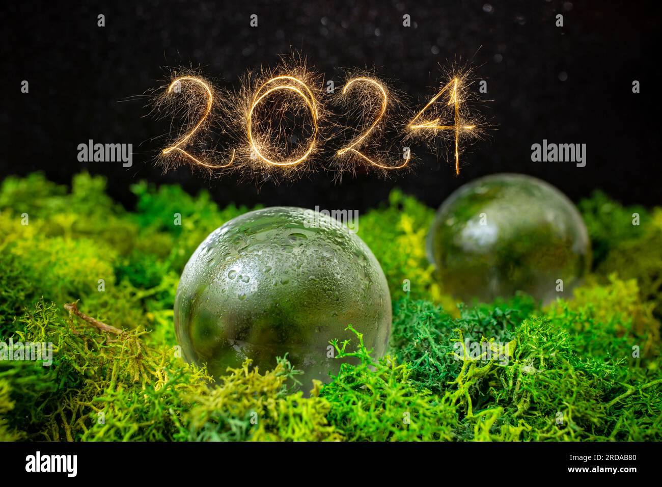 numero 2024 scritto con scintillanti dorati accanto a sfere di vetro su lichene verde con sfondo nero, messa a fuoco morbida in primo piano Foto Stock
