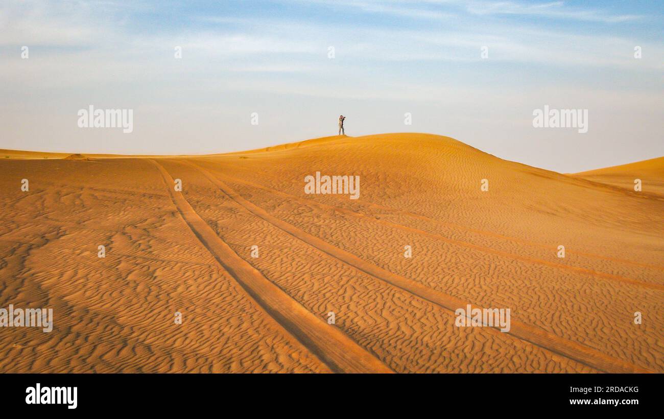 Paesaggio desertico con tracce dell'auto in primo piano e la figura del fotografo in lontananza, Dubai, Emirati Arabi Uniti Foto Stock