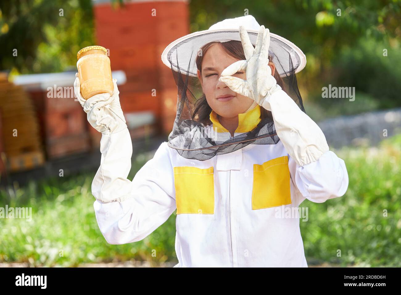 Ritratto di una ragazza che tiene in mano il vaso di miele e mostra il cartello OK che indossa una tuta protettiva all'apiario Foto Stock