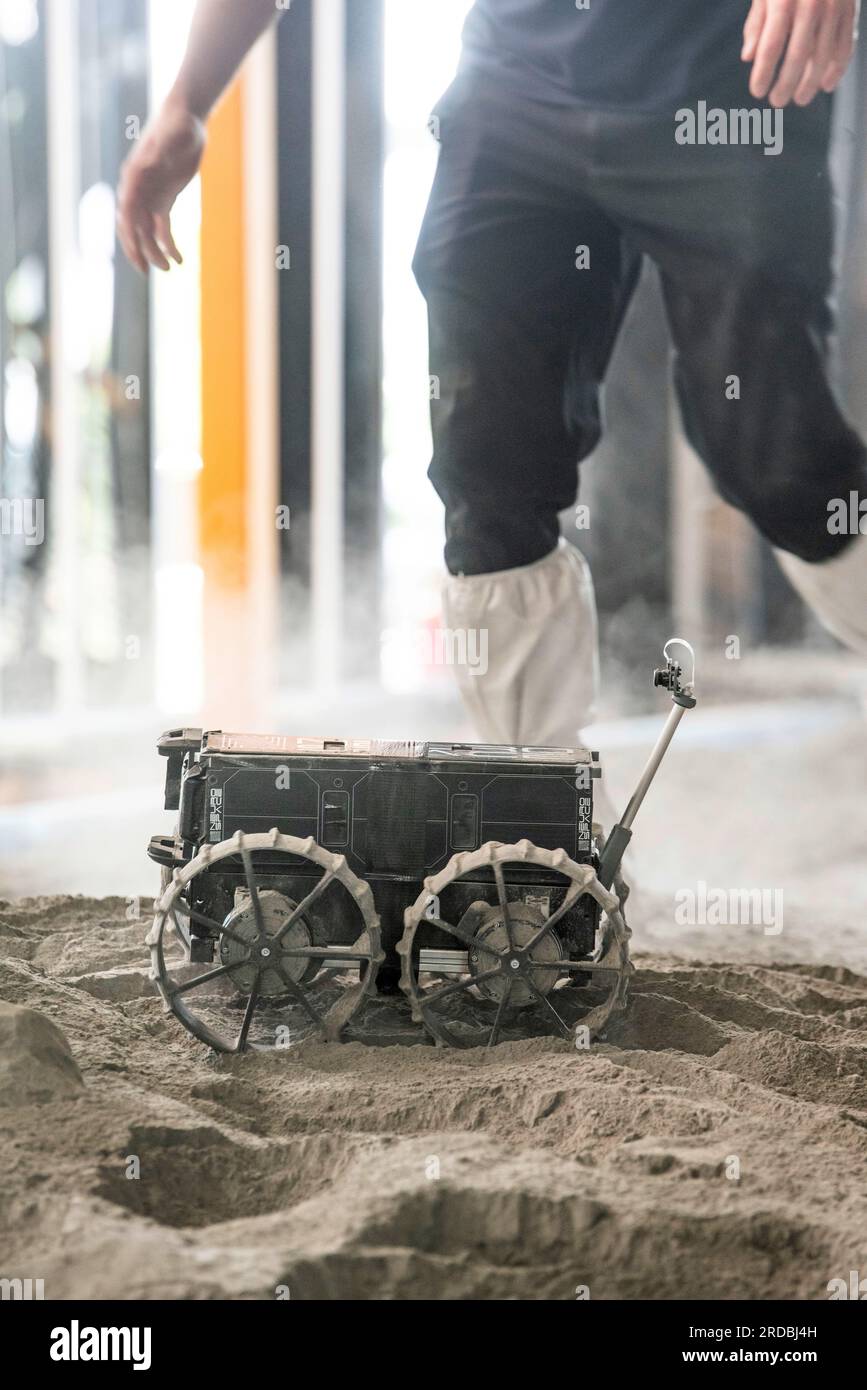 20 luglio 2023, Meclemburgo-Pomerania occidentale, Rostock-Laage: L'ingegnere di sviluppo Max Manthey della Neurospace GmbH accompagna il viaggio di un rover lunare su un campo di prova nel terminal aeroportuale dell'aeroporto di Rostock-Laage. Il letto lunare costruito artificialmente nel terminal dell'aeroporto è lungo circa 18 metri, largo cinque metri e pieno di "polvere lunare" sulla superficie. In occasione della giornata internazionale della Luna che celebra il primo atterraggio con equipaggio il 20 luglio 1969, i piccoli rover di prova stanno ancora arando attraverso un paesaggio lunare creato artificialmente all'aeroporto di Rostock-Laage. Foto: Frank Foto Stock