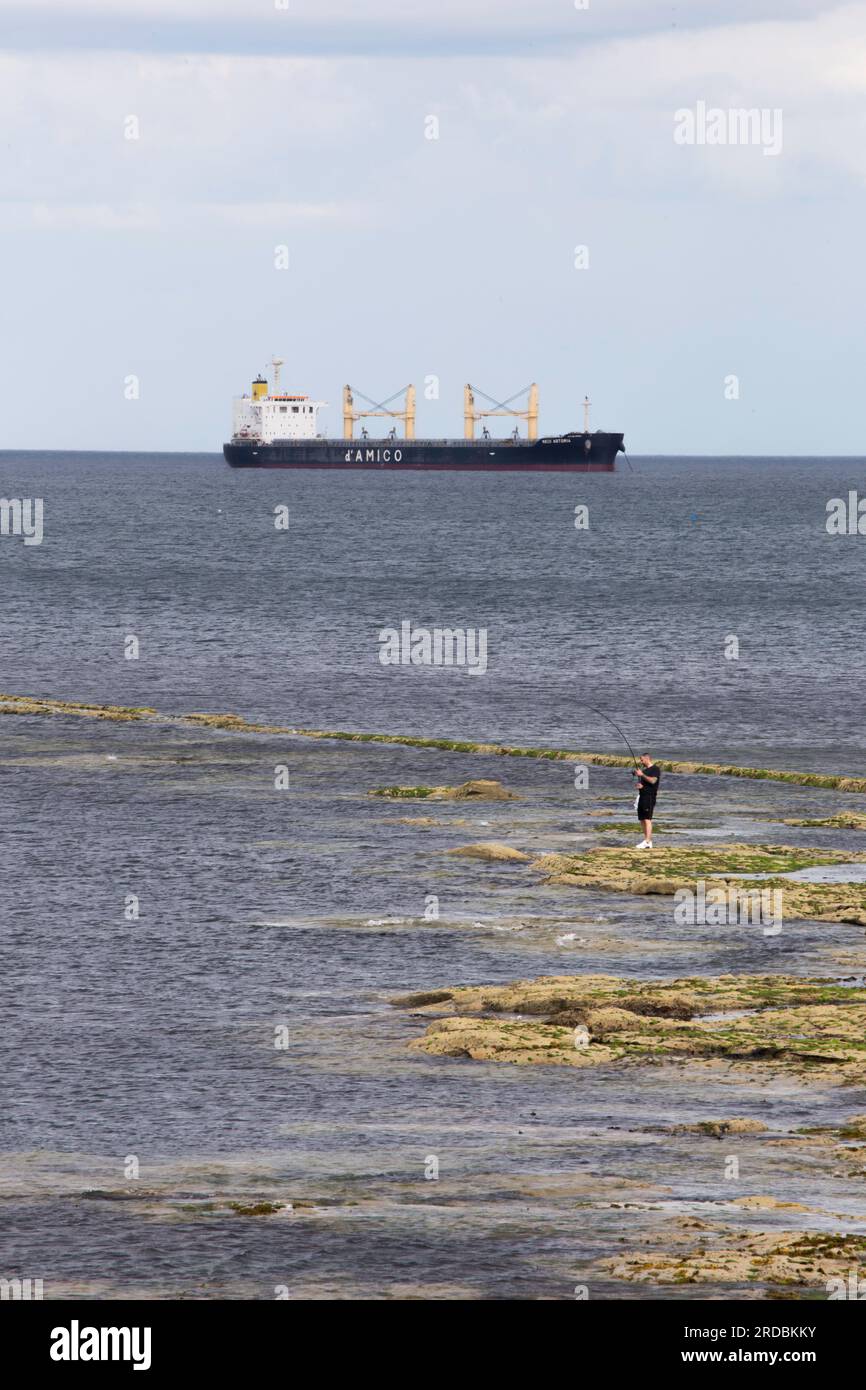 Un uomo solitario che pesca in mare con una grande nave sullo sfondo vicino a Whitley Bay Foto Stock