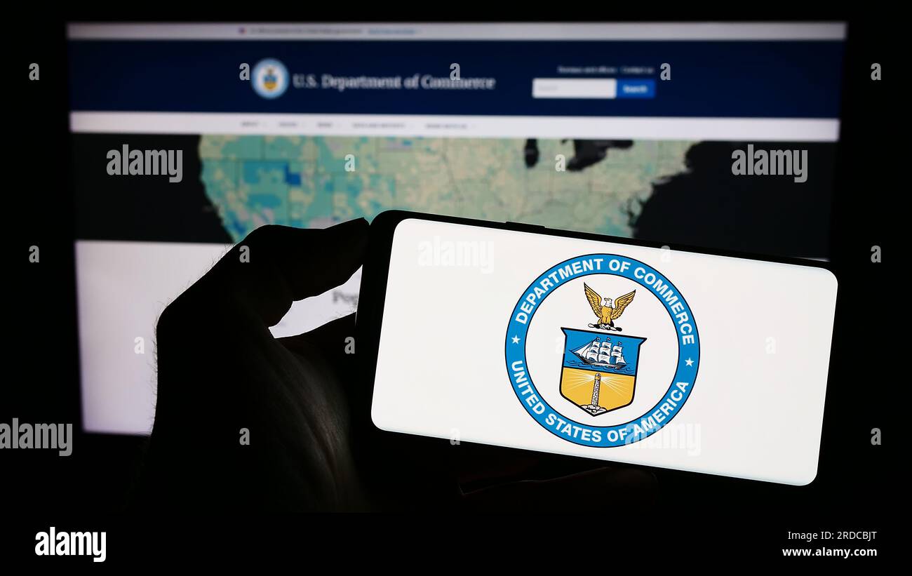 Persona in possesso di un cellulare con il sigillo del Dipartimento del commercio degli Stati Uniti (DOC) sullo schermo davanti alla pagina Web. Concentrarsi sul display del telefono. Foto Stock Persona in possesso di un cellulare con il sigillo del Dipartimento del commercio degli Stati Uniti (DOC) sullo schermo davanti alla pagina Web. Concentrarsi sul display del telefono. Foto Stock