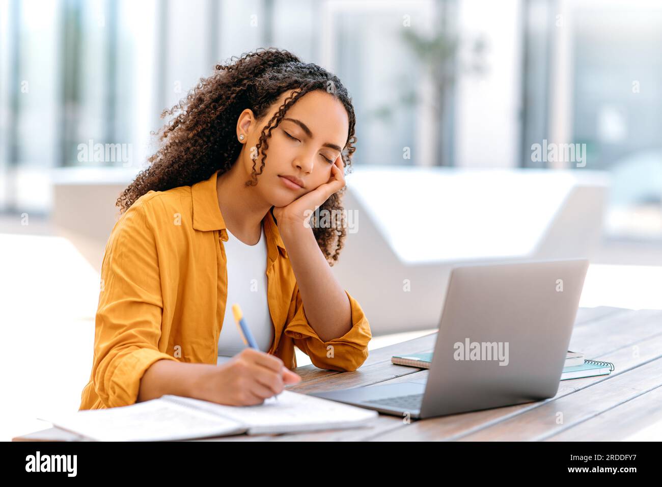 Dormi al lavoro o allo studio. Stanca ragazza brasiliana dai capelli ricci o latina, siede all'aperto con un notebook, si addormenta mentre studia o lavora online, si sente stanca e manca il sonno, mentre dorme a occhi chiusi Foto Stock