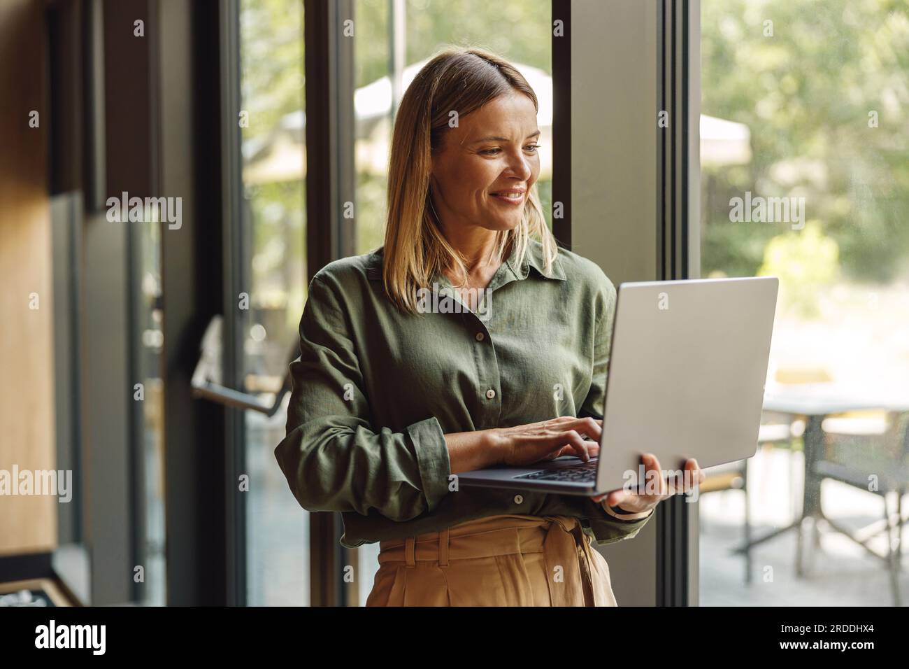 Donna d'affari sorridente che lavora su un computer portatile in un accogliente ufficio in piedi vicino alle finestre Foto Stock