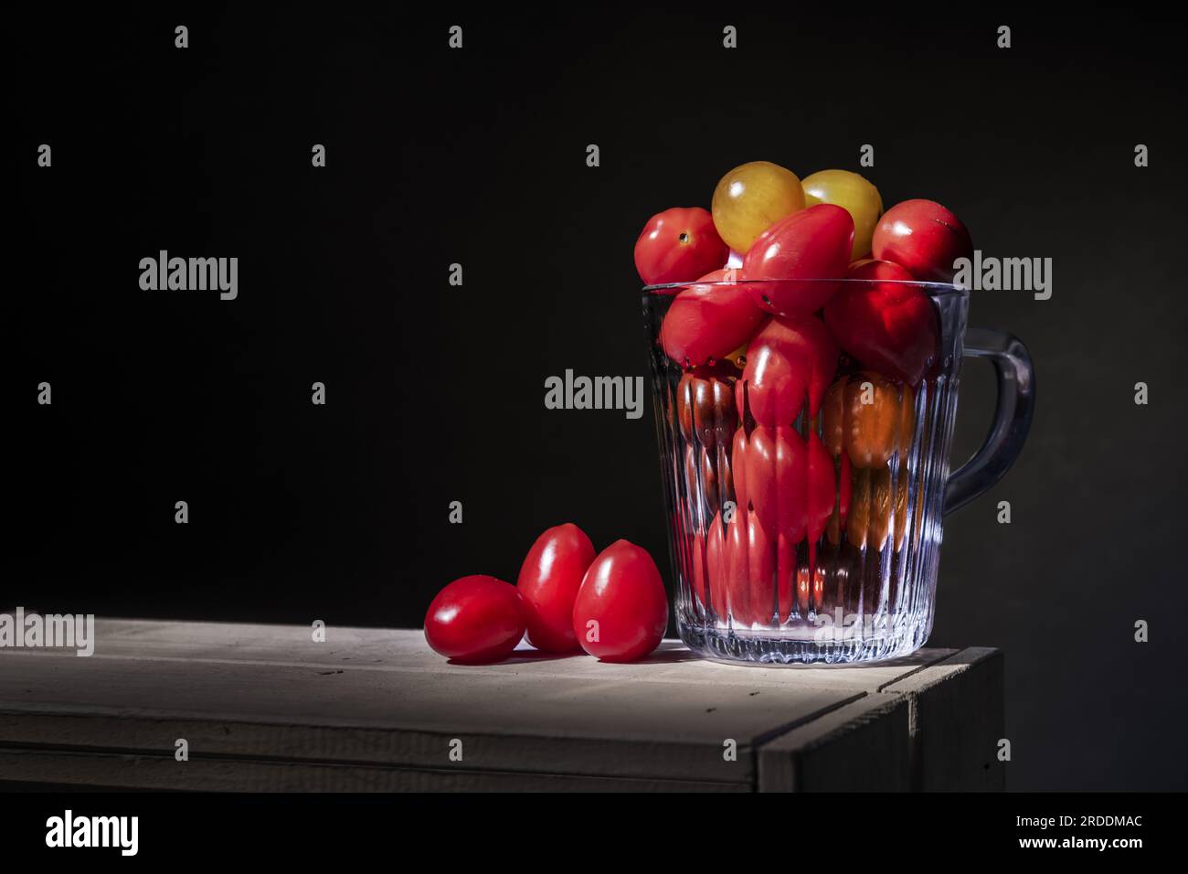 Un piccolo bicchiere con manico in vetro trasparente pieno di pomodori ciliegini su un tavolo di legno bianco Foto Stock