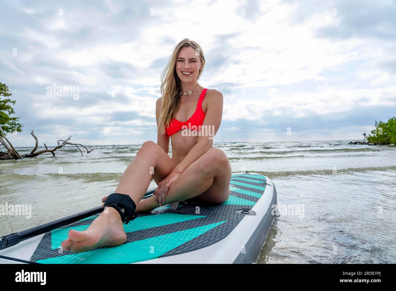 Un bel modello biondo gode di una giornata estiva mentre si prepara a navigare sull'oceano con la sua tavola da pagaia Foto Stock