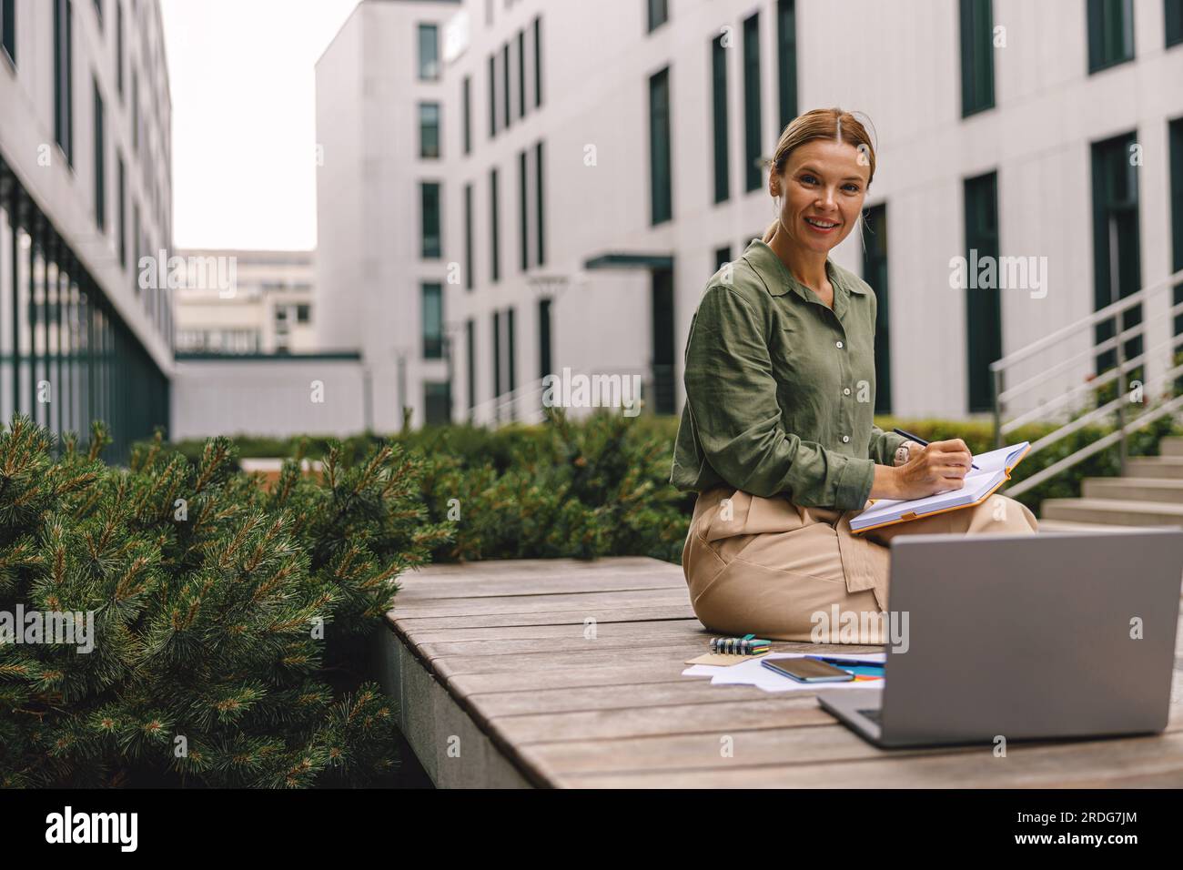 Analista donna che lavora con documenti e utilizza un notebook seduto all'esterno su un edificio moderno Foto Stock