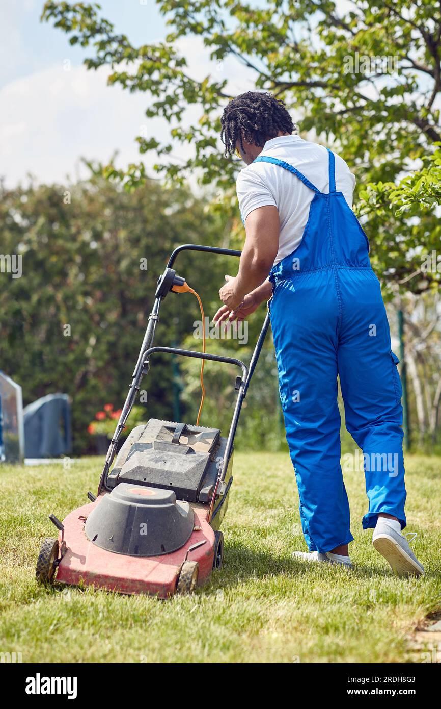 Un uomo afro-americano gestisce con sicurezza un rasaerba. Con un'espressione focalizzata e una presa salda sulle impugnature, fa scivolare il rasaerba senza sforzo Foto Stock