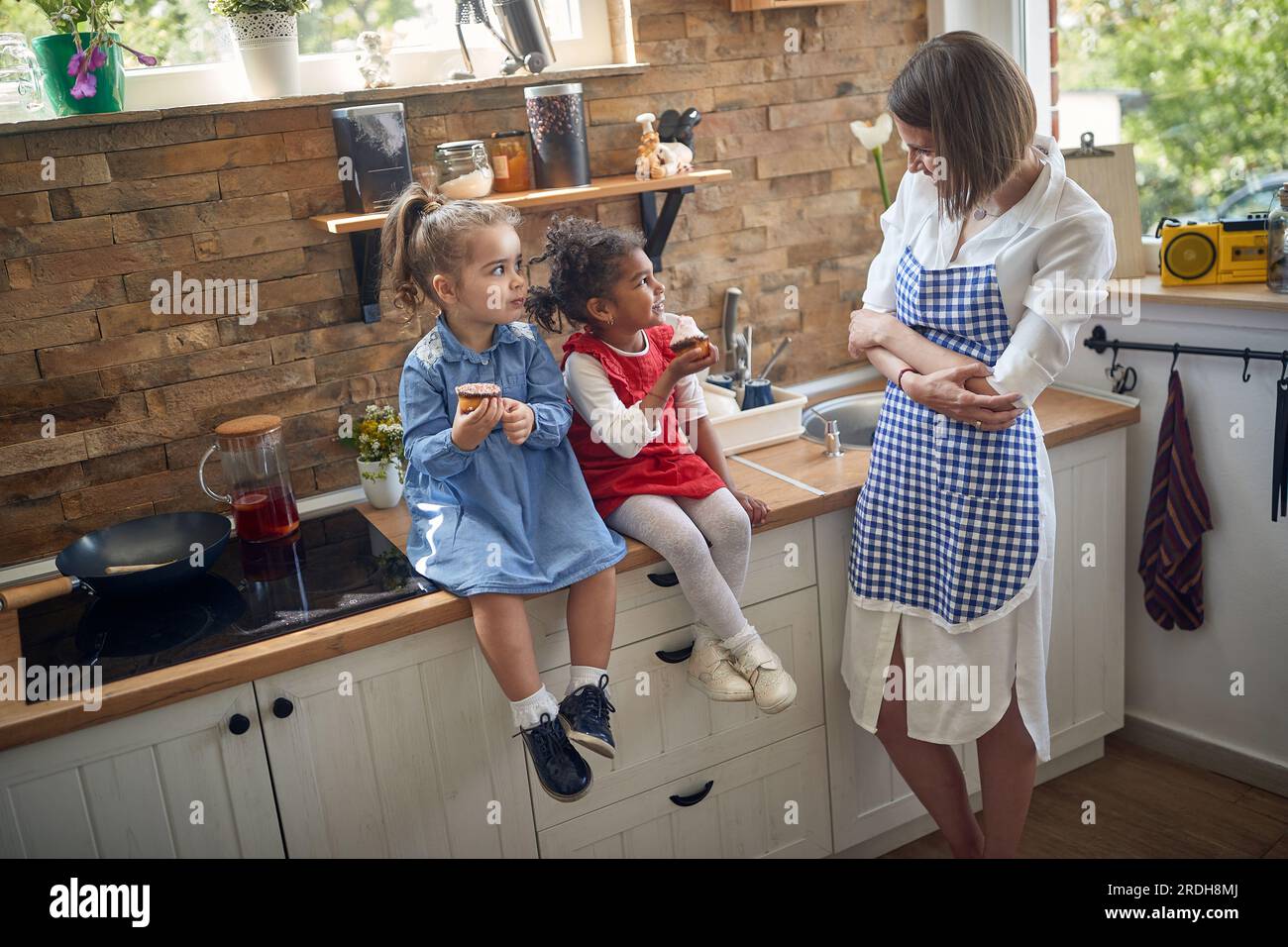 Due ragazze, sedute insieme in cucina, assaporando deliziosi muffin. In piedi accanto a loro, la loro madre travolge con orgoglio e gioia, il suo sorriso a testa Foto Stock