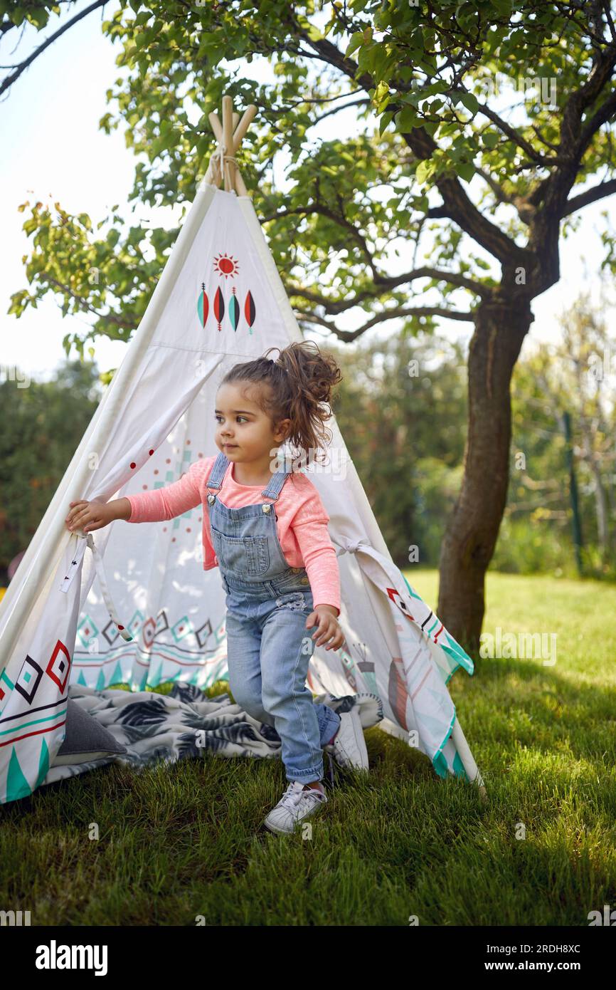 Adorabile bambina che gioca all'aperto nel cortile, uscendo da una tenda bohémien. Casa, stile di vita, concetto di vacanza. Foto Stock
