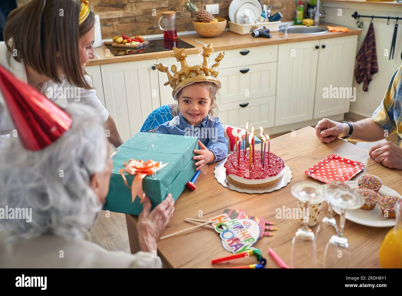 Felice bambina con una corona gonfiabile che festeggia il suo compleanno,  seduta al tavolo con gli ospiti, ricevendo regali da sua nonna. A casa, fam  Foto stock - Alamy