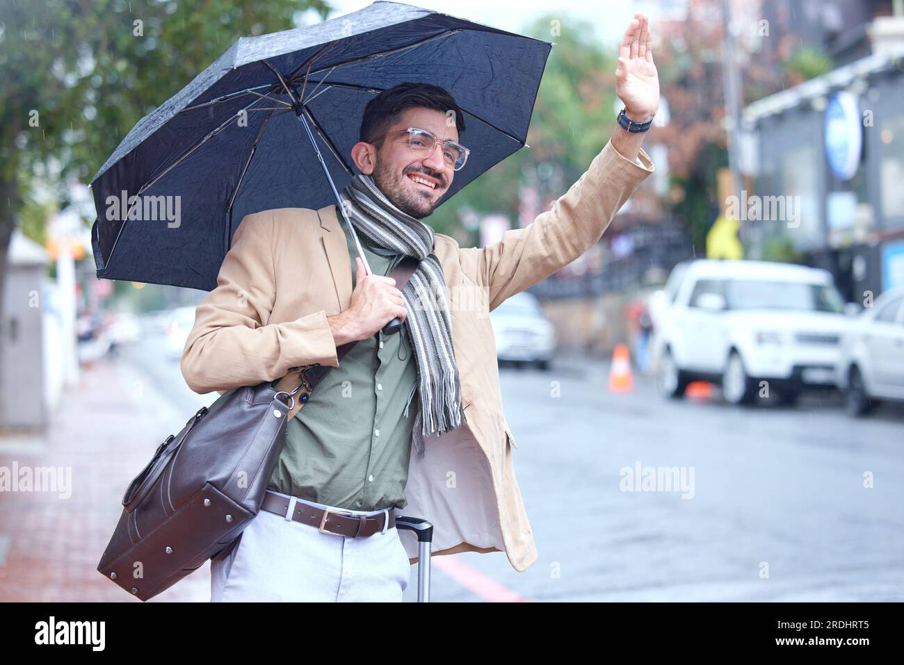 Uomo felice, ombrellone e taxi in città per viaggiare, sollevare o prelevare e aspettare sul marciapiede della strada sotto la pioggia. Uomo che salta la mano per segnale, viaggio o. Foto Stock