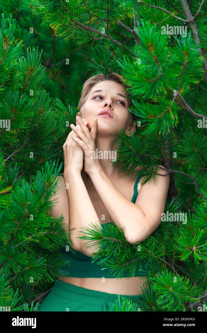 Timida e attraente donna bionda posa sullo sfondo di un arbusto sempreverde di Stone Pine giapponese. Elegante modello europeo vestito con top corto verde Foto Stock