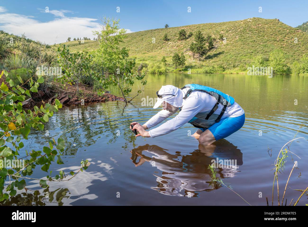Un uomo anziano che indossa un giubbotto salvagente e un cappello da sole sta scattando foto con un telefono in acqua vicino alla riva, il lago artificiale Horsetooth in Colorado Foto Stock