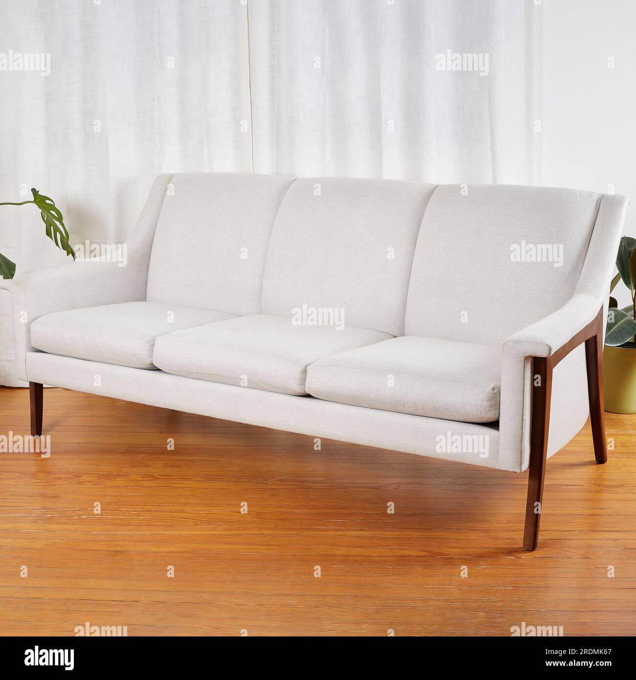 Divano vintage bianco. Elegante divano moderno della metà del secolo con gambe in legno scultoreo. Scena interna con tende bianche e pavimento in legno. Foto Stock