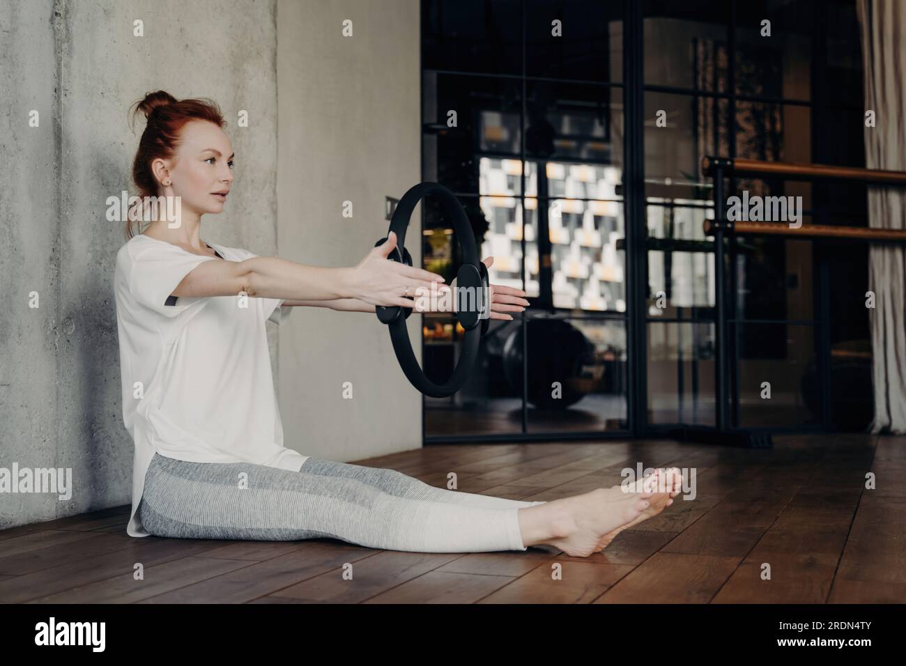Donna sportiva concentrata, con t-shirt bianca e leggings grigi, siede in una sala fitness e fa esercizi con il braccio con l'anello Pilates. Gambe allungate. Foto Stock