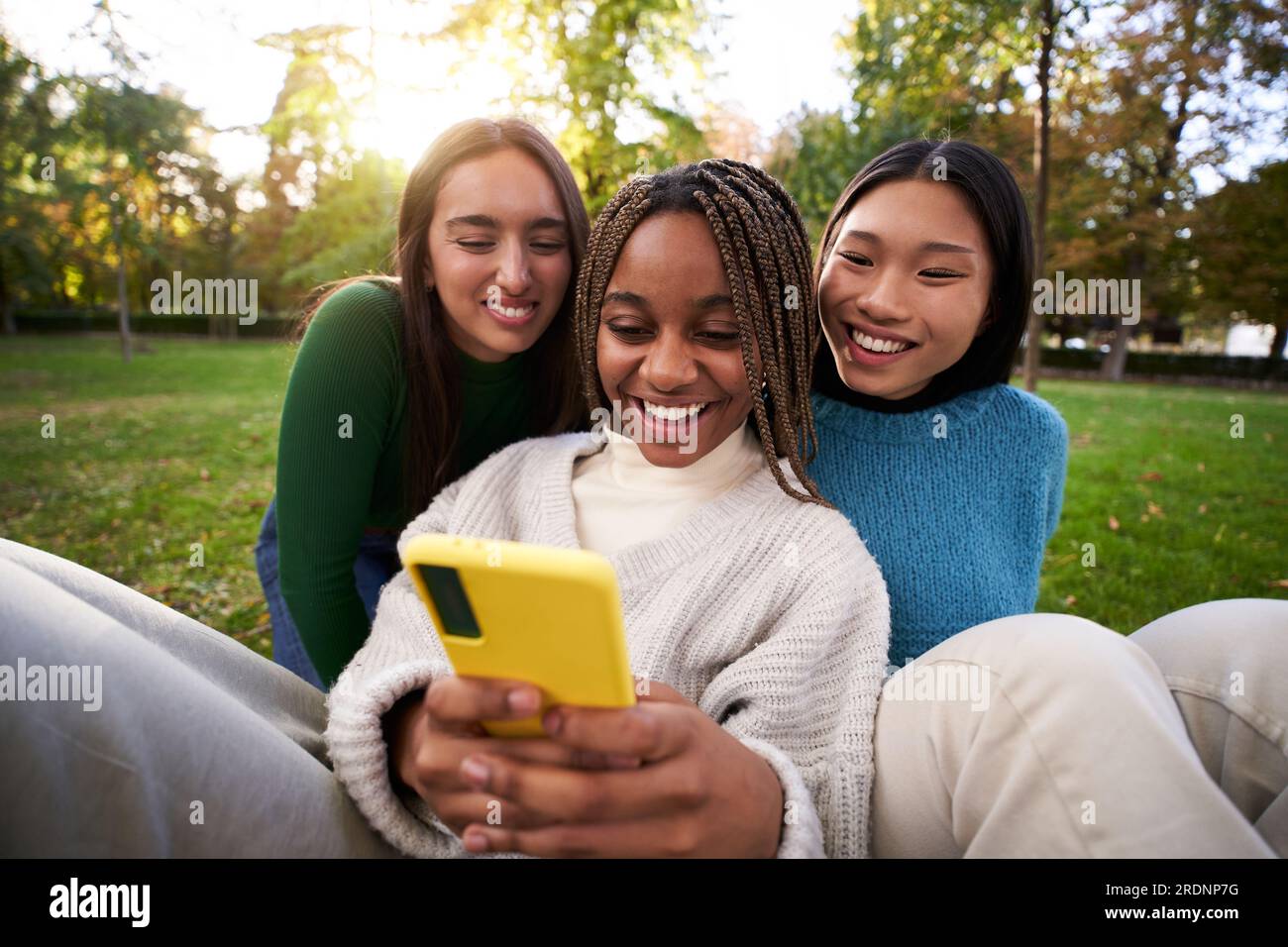 Tre giovani donne multirazziali che usano un cellulare sorridente nel parco. Persone Millennial connesse online. Foto Stock