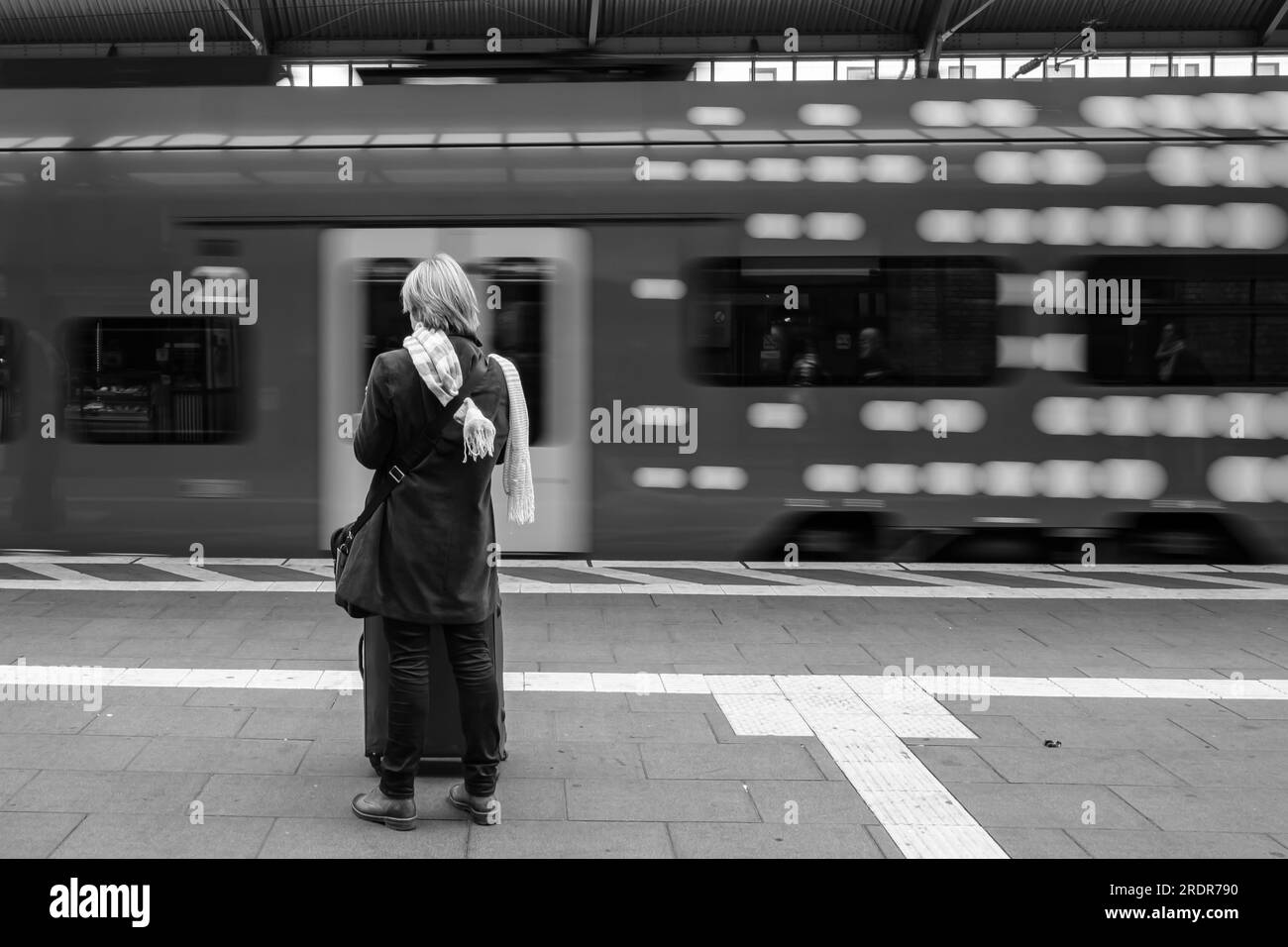 Veduta di una sola donna in piedi alla stazione ferroviaria centrale di Bonn in Germania e di un treno che lascia il binario in bianco e nero Foto Stock