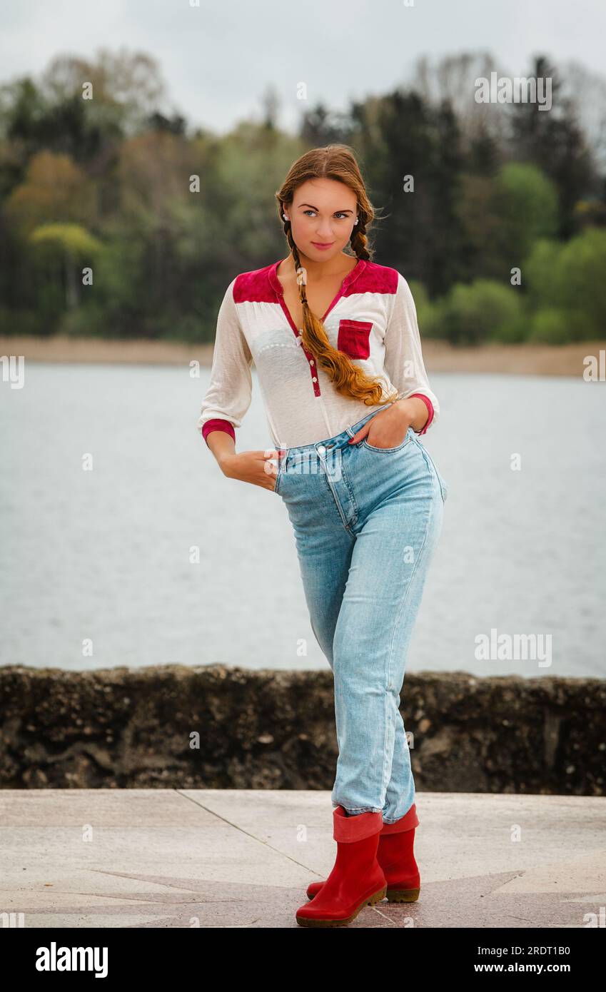 Giovane donna dai capelli lunghi sulla riva del lago con jeans e stivali in gomma. Foto Stock