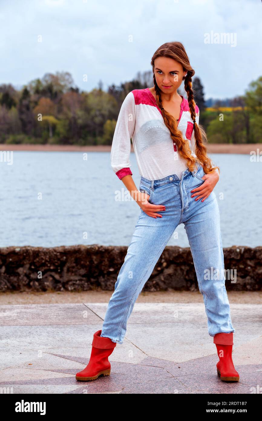 Giovane donna con i capelli lunghi sulla riva del lago in jeans e stivali in gomma in posa modello Foto Stock