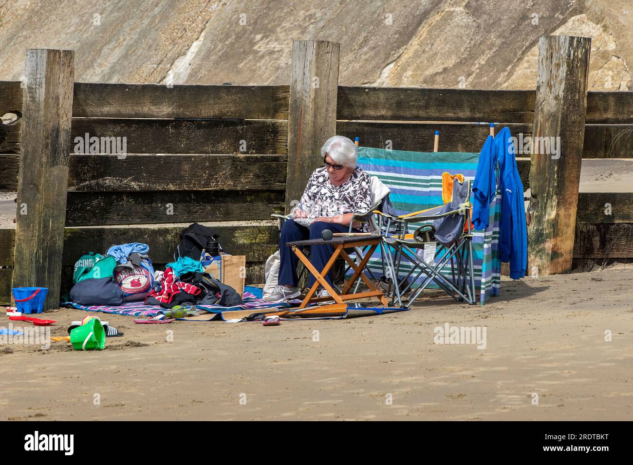signora anziana che si prende la calma sedersi su una sedia dietro un vento sulla spiaggia di sandown sull'isola di wight, una donna matura britannica seduta su una spiaggia. Foto Stock