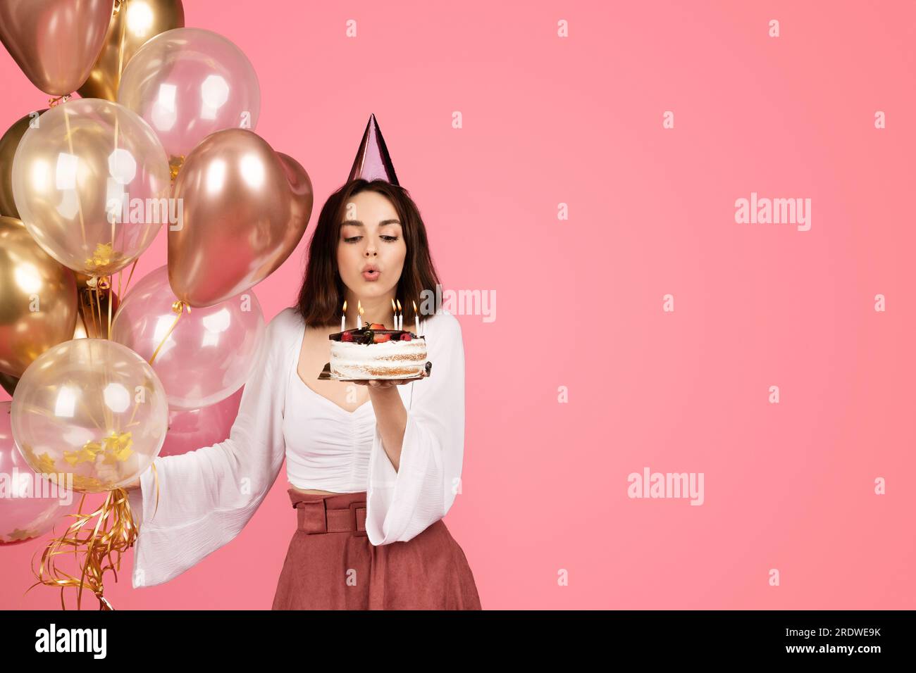 Allegra donna bruna caucasica millenaria con un cappello e molti palloncini sprigiona le candele sulla torta Foto Stock