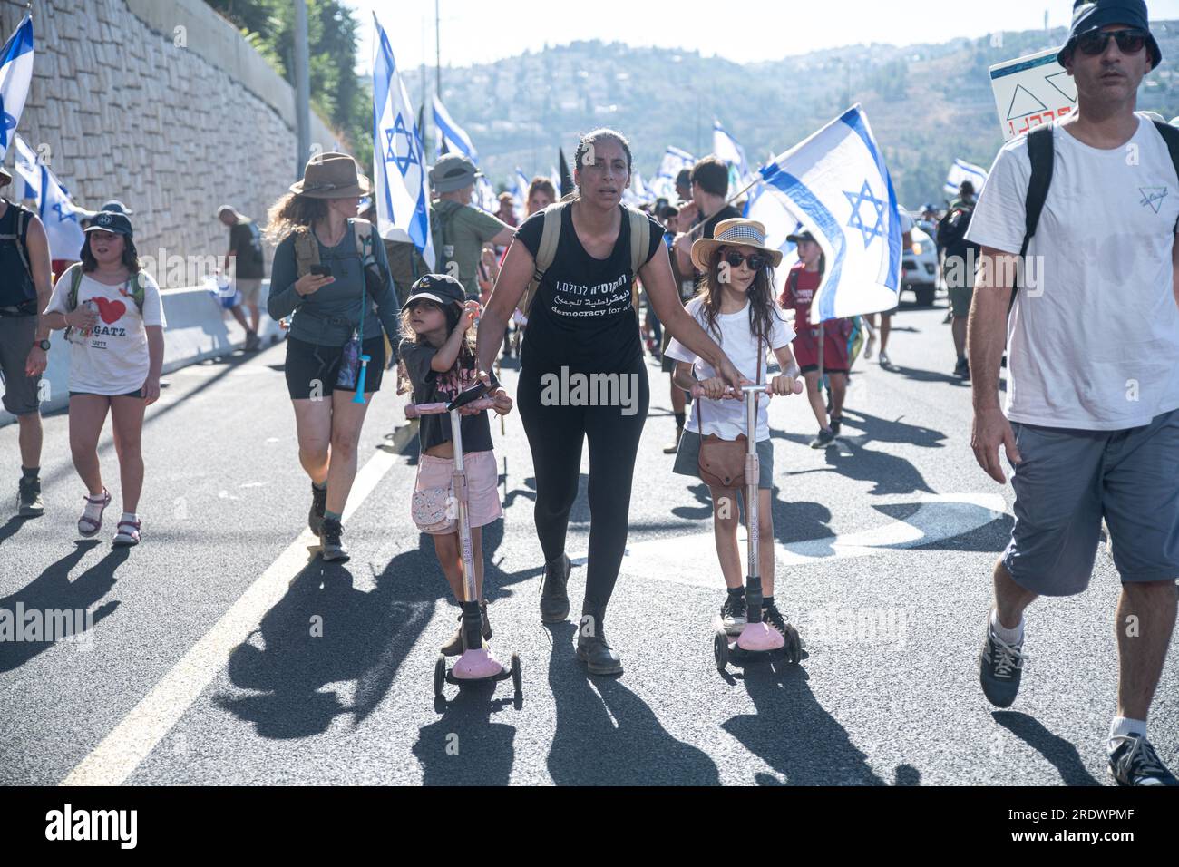 Un protestore contro la riforma spinge le sue figlie a scooters mentre scalano la strada d'ingresso a Gerusalemme durante la manifestazione. Decine di migliaia di manifestanti contro la riforma sventolano la bandiera israeliana mentre si arrampicano sulla strada d'ingresso a Gerusalemme, terminando una marcia di 4 giorni a partire da Tel Aviv in una manifestazione contro la legislazione in corso di revisione legale. Foto Stock