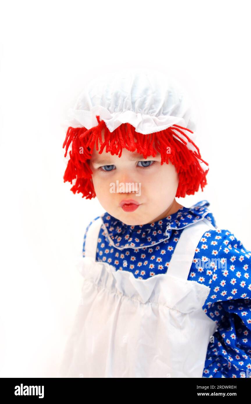 La bambina arrabbiata e incrociata è vestita con un costume da bambola da straccio. I suoi capelli sono in filato rosso ricoperti da un cappello bianco per mop. Foto Stock