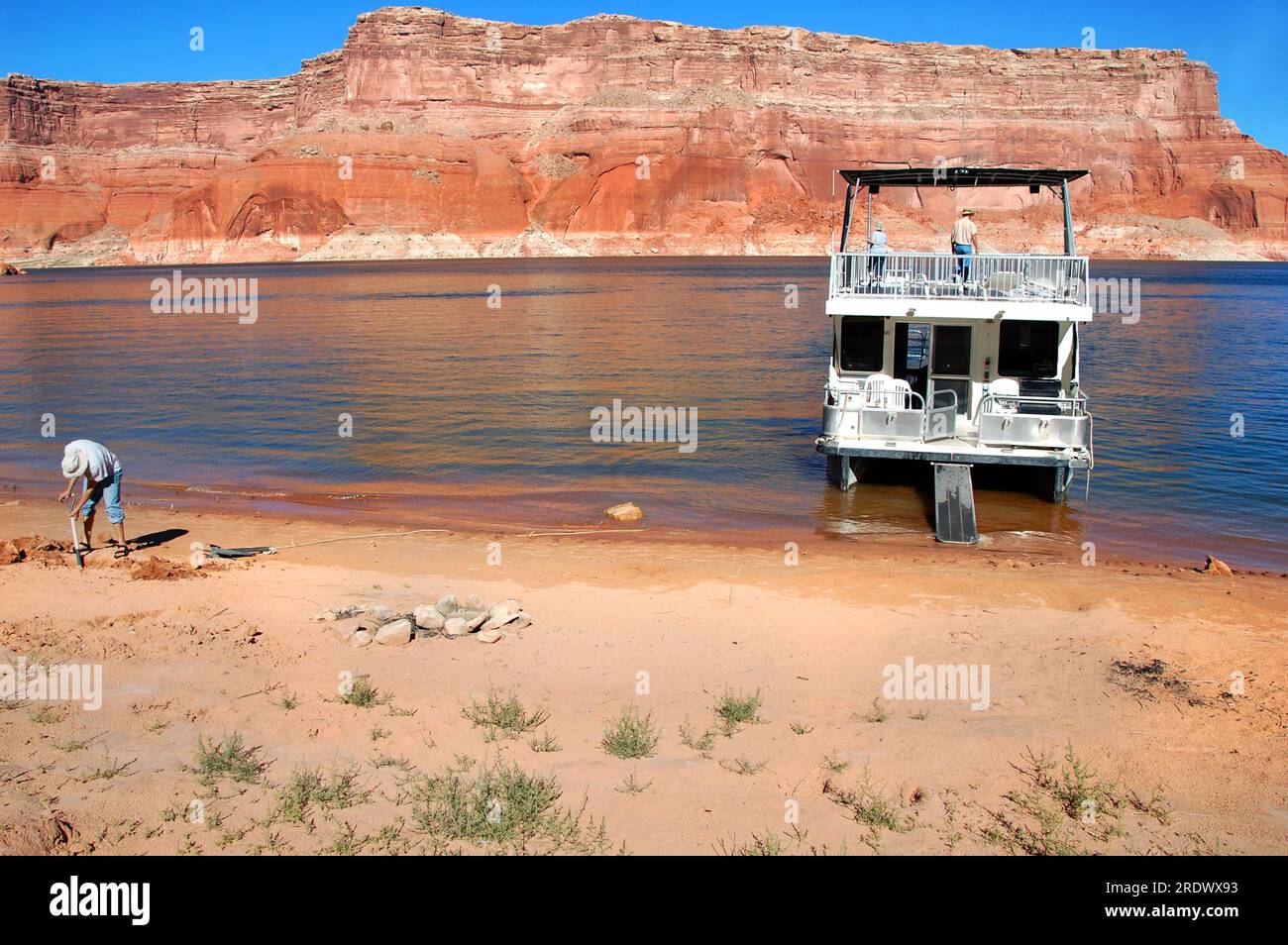 I vacanzieri preparano la casa galleggiante per il campeggio notturno. L'uomo scava sulla riva per seppellire l'ancora. Le scogliere di arenaria rossa si affacciano sulle acque blu del lago Powell Foto Stock