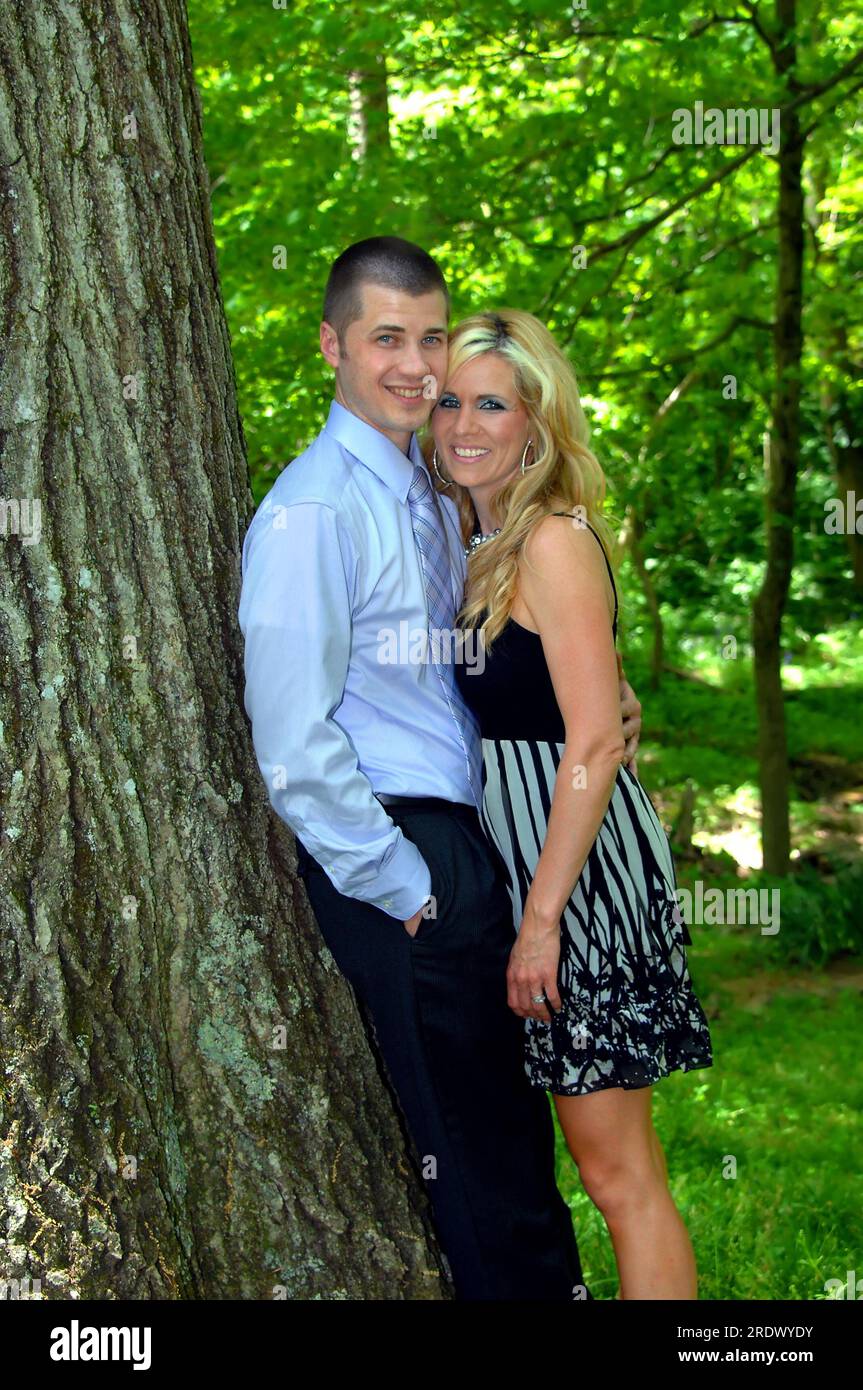 Giovane coppia appoggiata contro un tronco d'albero e vicina l'una all'altra. Sorridono e guardano la fotocamera. Entrambi indossano abiti da dressy. Foto Stock