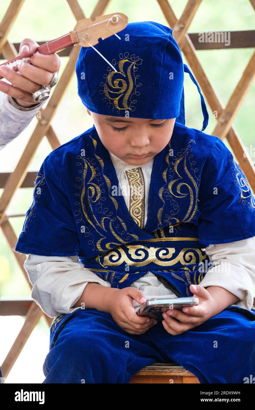 Kazakistan, Huns Ethno Village. Giovane ragazzo kazako in abito tradizionale guardando il suo cellulare. Foto Stock