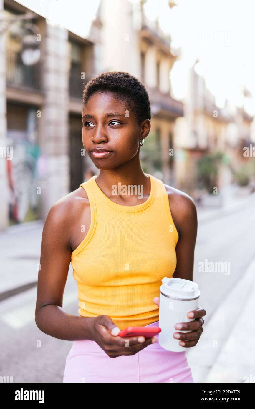 premurosa donna africana in piedi per strada e guardando lontano. Ha in mano una tazza da caffè riutilizzabile e uno smartphone. Foto Stock