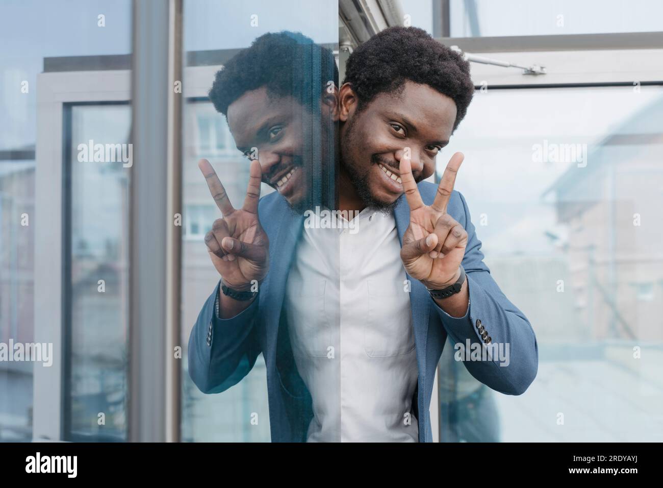Un uomo d'affari sorridente che mostra un segno di pace che sbircia dalla finestra di vetro dell'ufficio Foto Stock