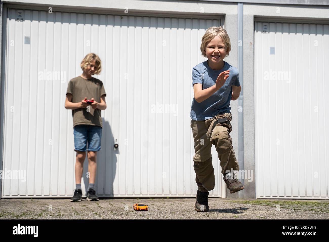 Happy boy che balla e fratello che gioca con un'auto telecomandata nel cortile fuori dal garage Foto Stock