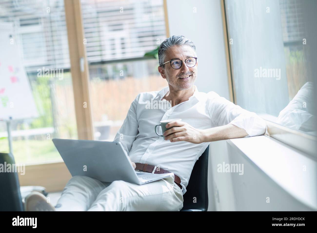 Un uomo d'affari premuroso che tiene una tazza da caffè e un computer portatile che guarda fuori dalla finestra dell'ufficio Foto Stock