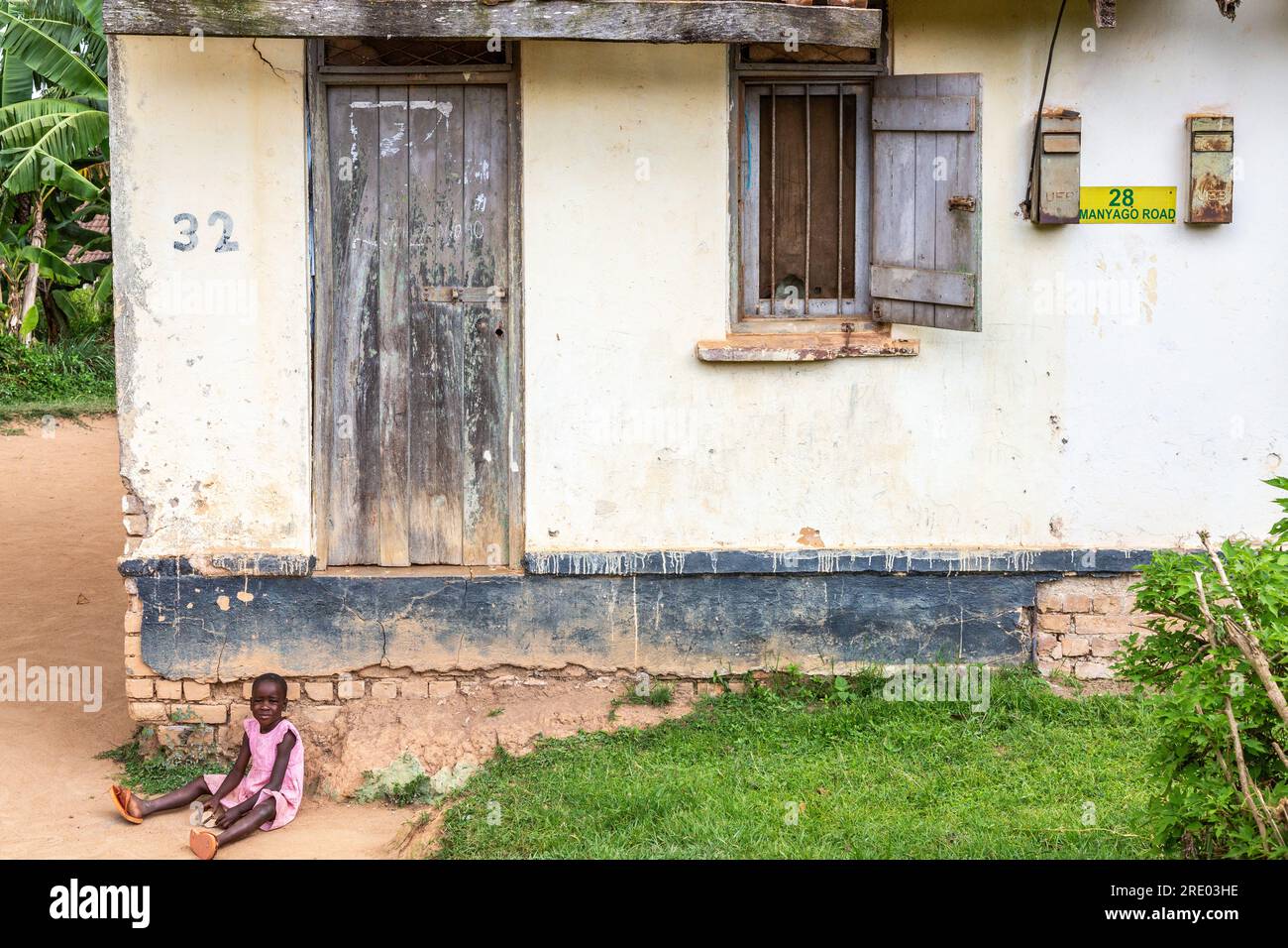 Casa residenziale molto modesta in una strada a Entebbe, Uganda. Giovane ragazza vestita di rosa seduta alla soglia della porta. Foto Stock