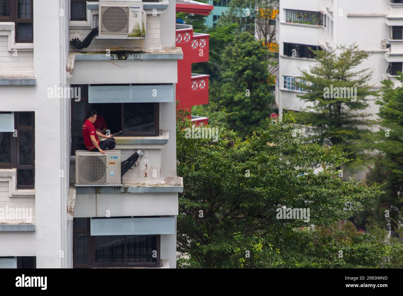 Un tecnico è seduto sull'unità aria condizionata in un edificio alto che esegue riparazioni a/C e dispone di imbracature di sicurezza per evitare cadute dall'alto. Foto Stock