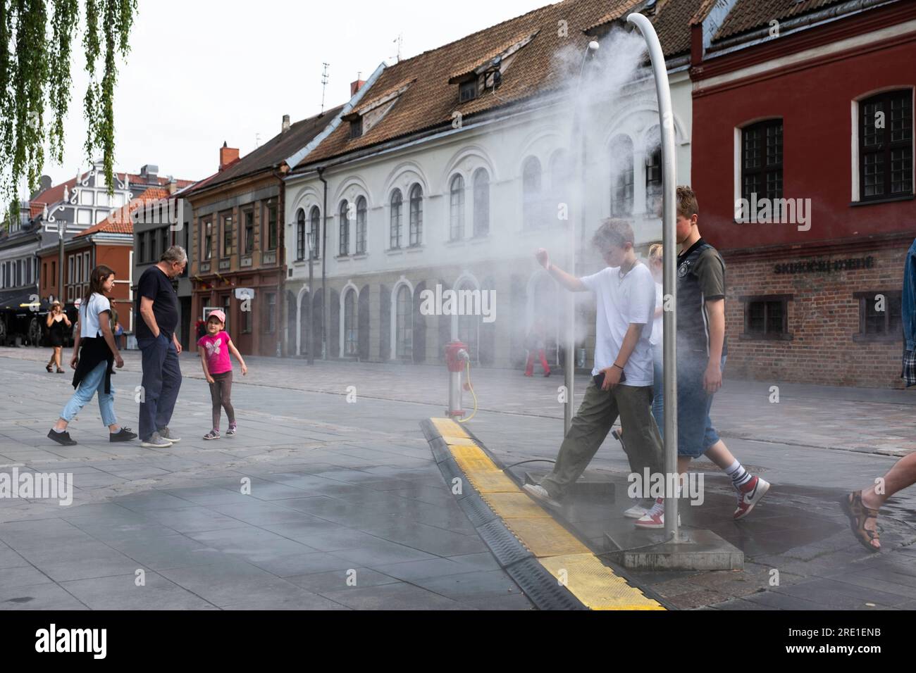I ragazzi camminano attraverso un arco metallico di un irrigatore ad acqua per rinfrescarsi a causa dell'appannamento, in una piazza a Kaunas. Pericolo di contaminazione da Legionella Foto Stock